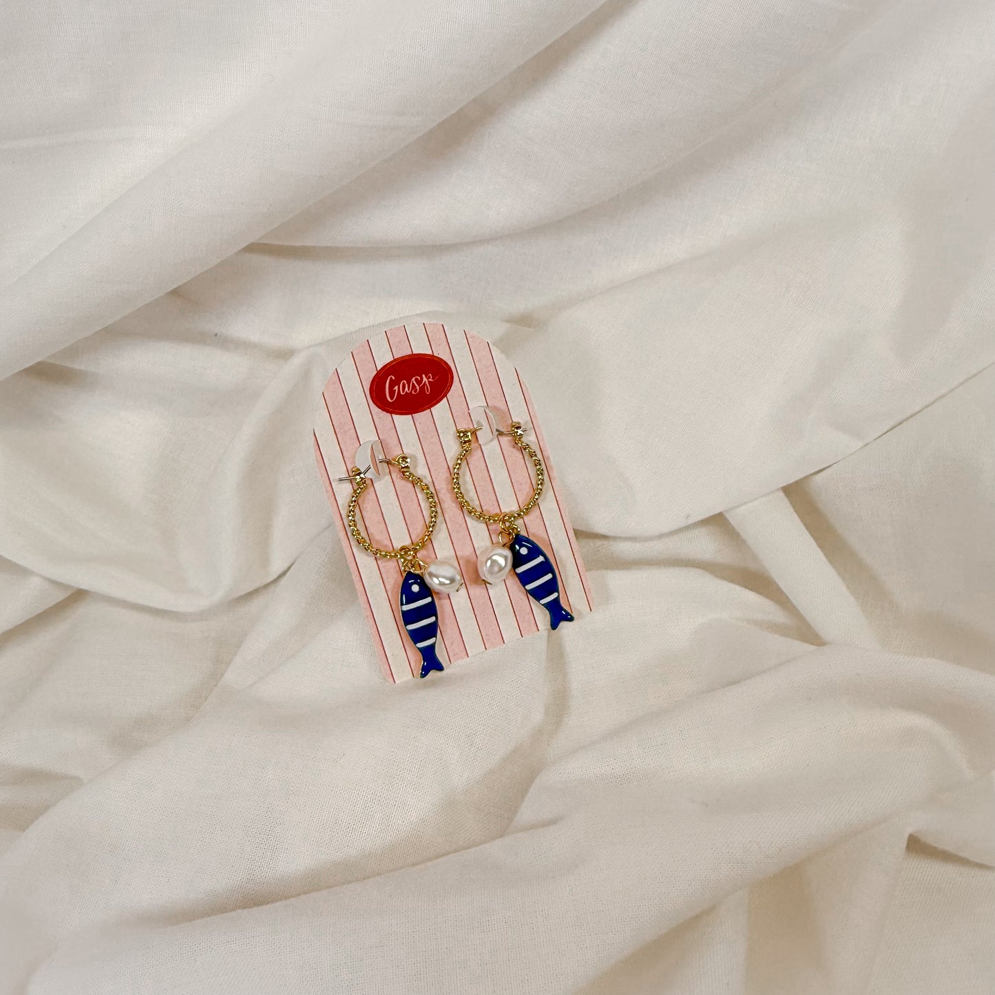 Gold hoop earrings with blue ceramic fish charm, sitting on a white fabric background
