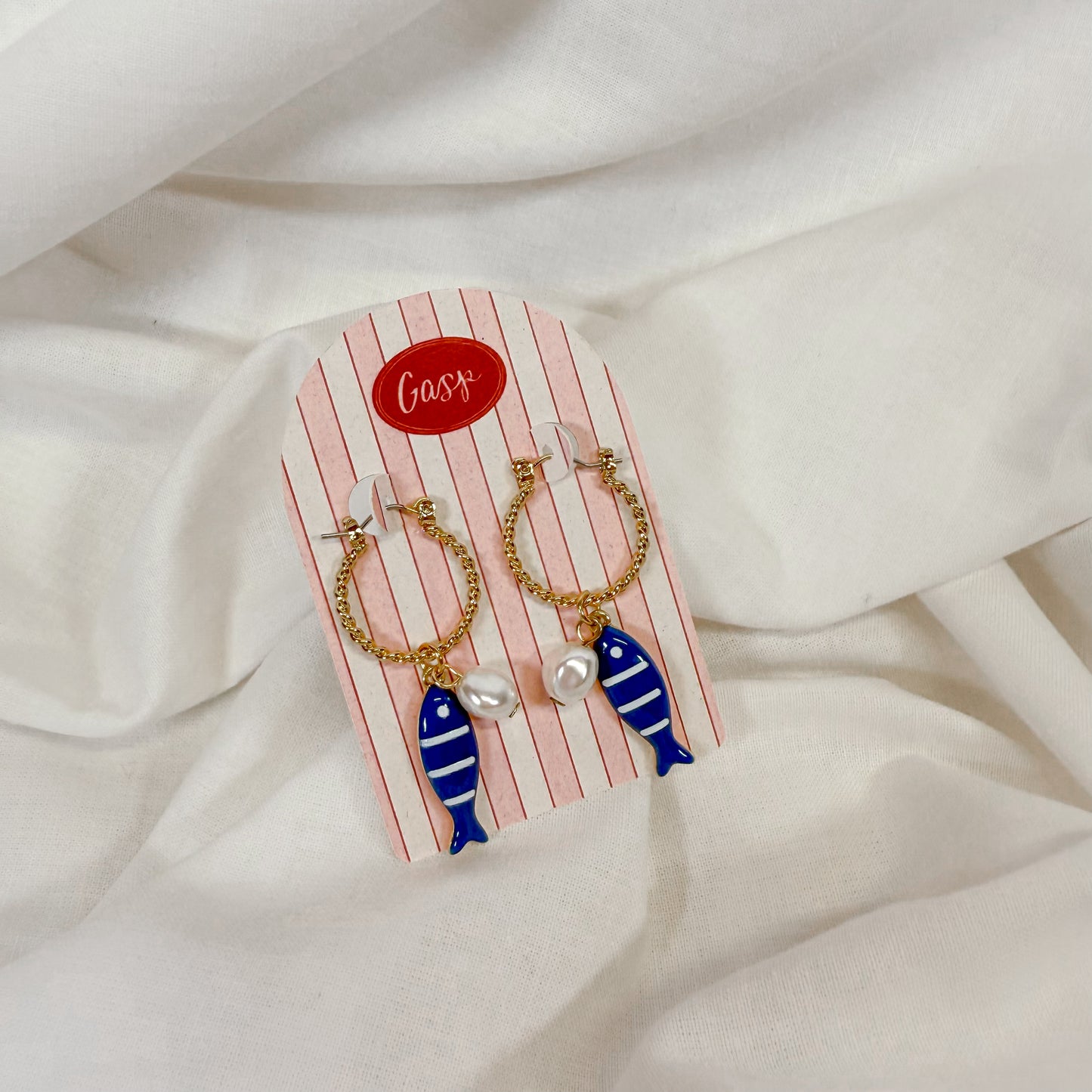 Earrings with fish and pearl design on a striped card against a white fabric background