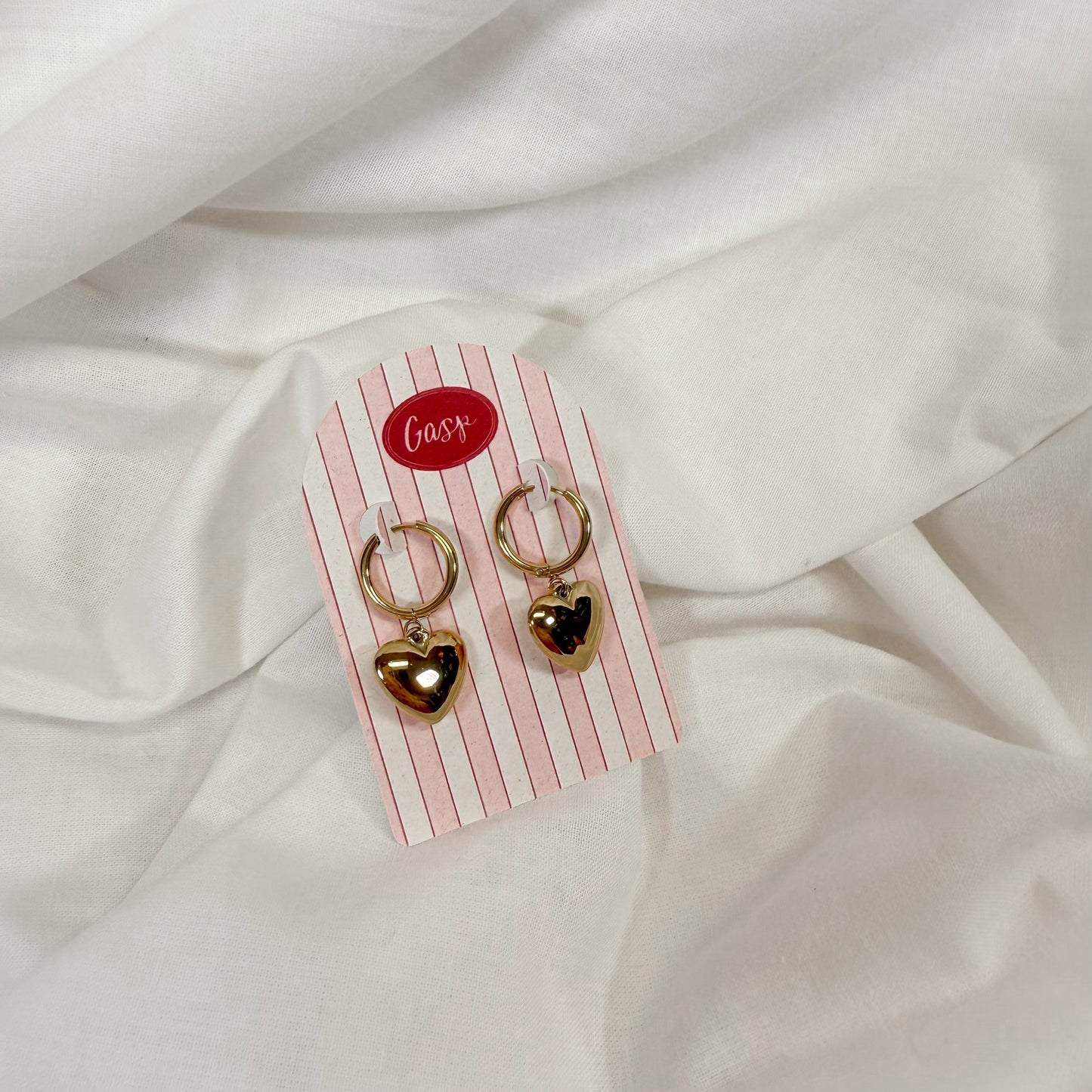 Gold heart-shaped dangly earrings on a striped card with 'Gasp' branding, placed on a white fabric background.