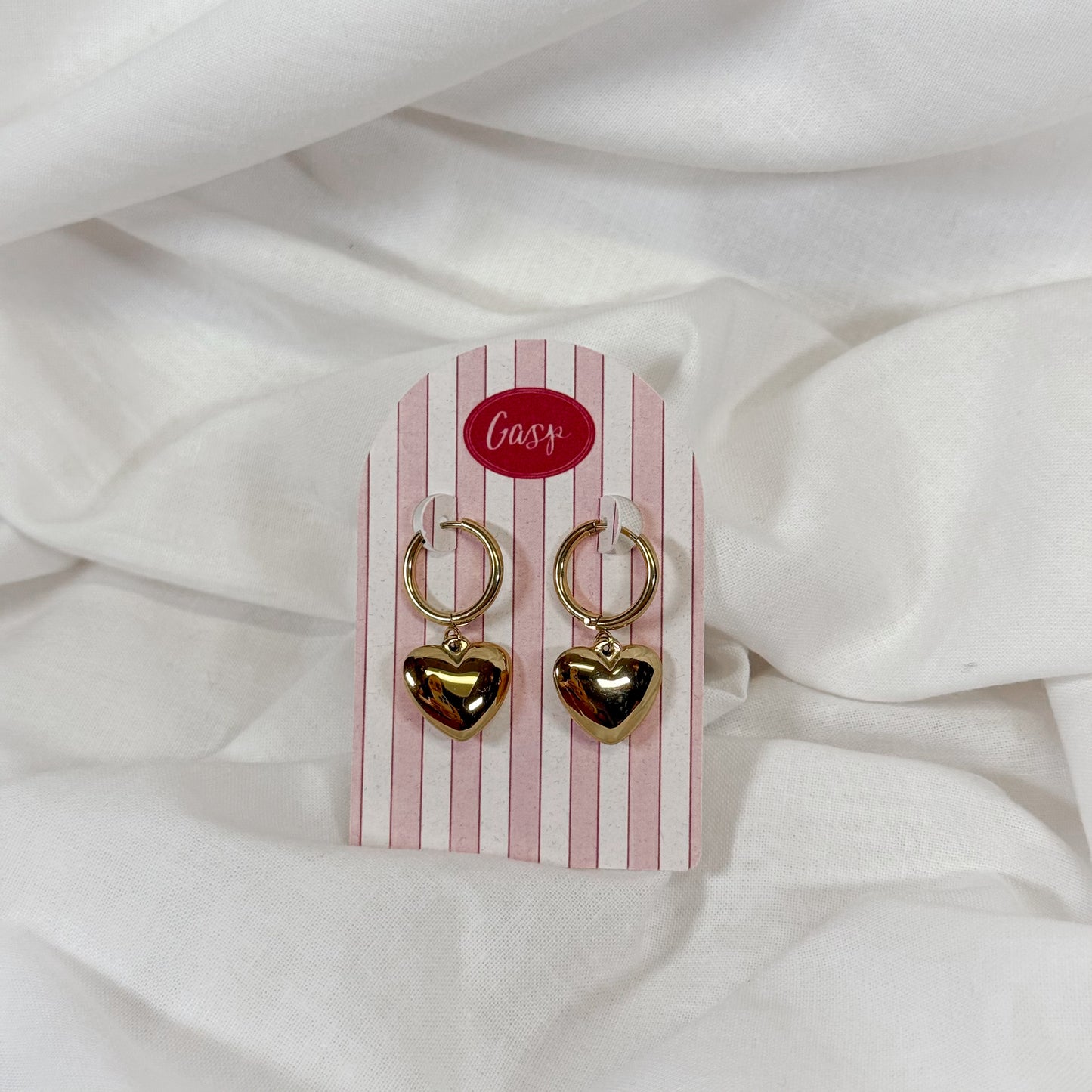 Puffy gold heart-shaped earrings on a pink and white striped card with 'Gasp' brand name, against a white fabric background.