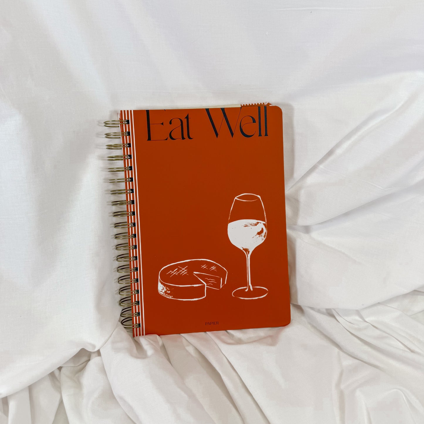 Notebook titled 'Eat Well' with a wine glass and cheese illustration on a white fabric background