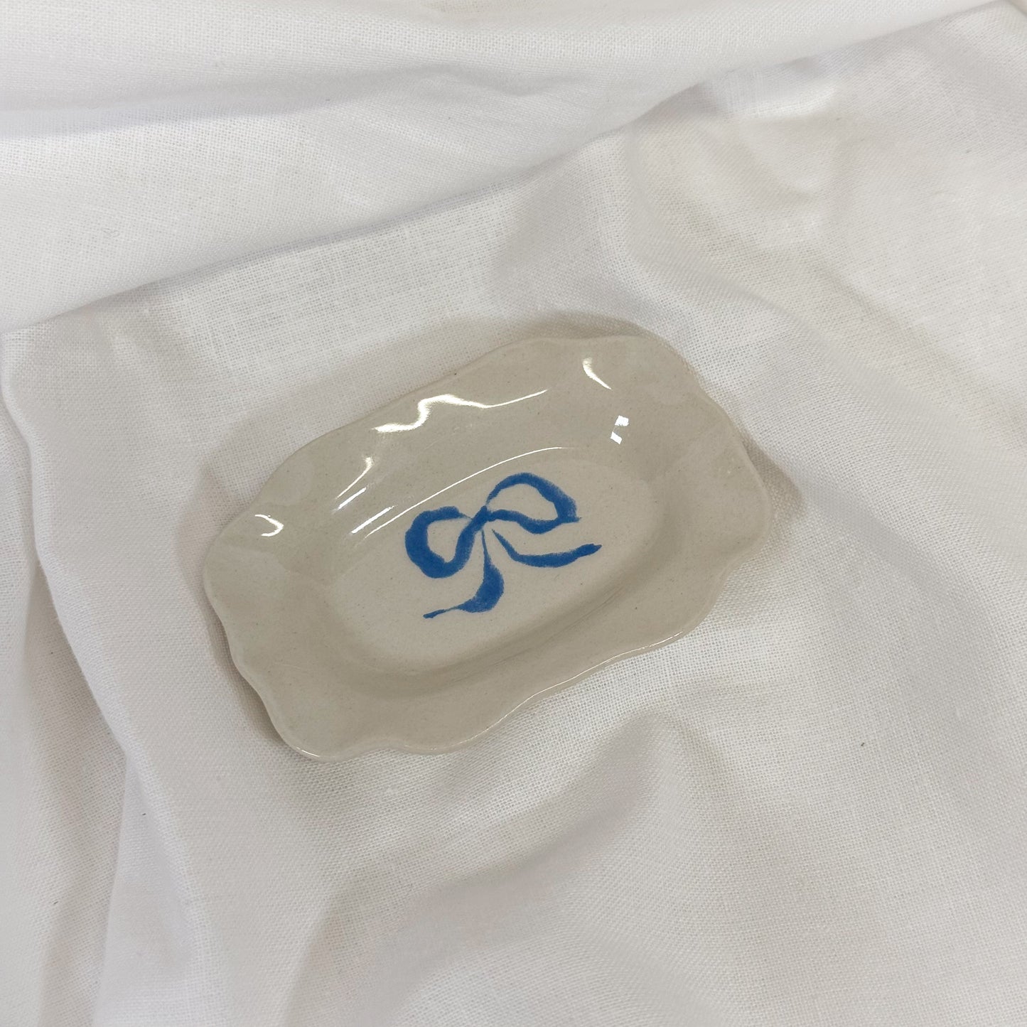 Cream colored jewelry dish with a blue ribbon bow on a white fabric background