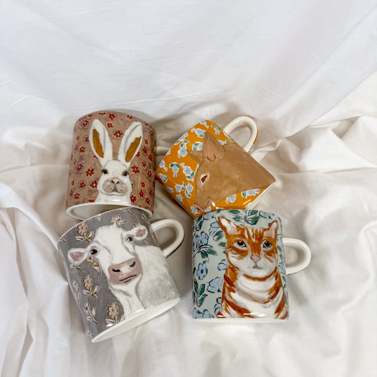 Four ceramic mugs with animal illustrations on a white fabric background