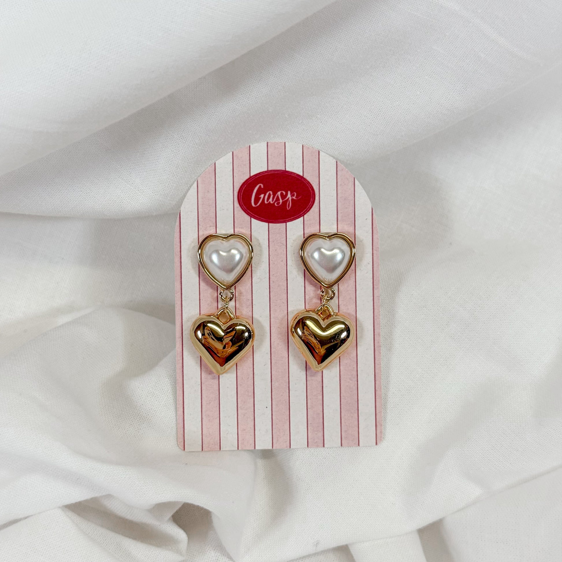 Heart-shaped pearl earrings on a striped card with 'Gasp' branding against a white fabric background