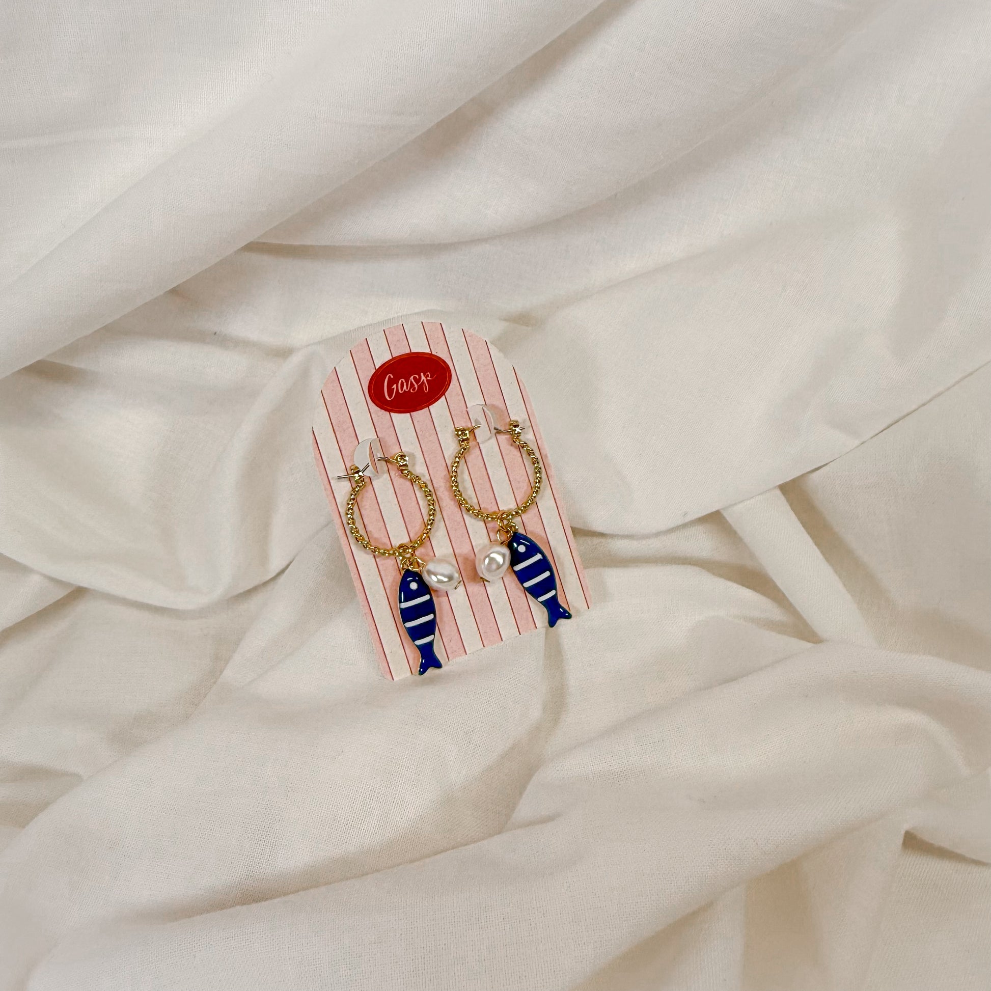 Gold hoop earrings with blue ceramic fish charm, sitting on a white fabric background