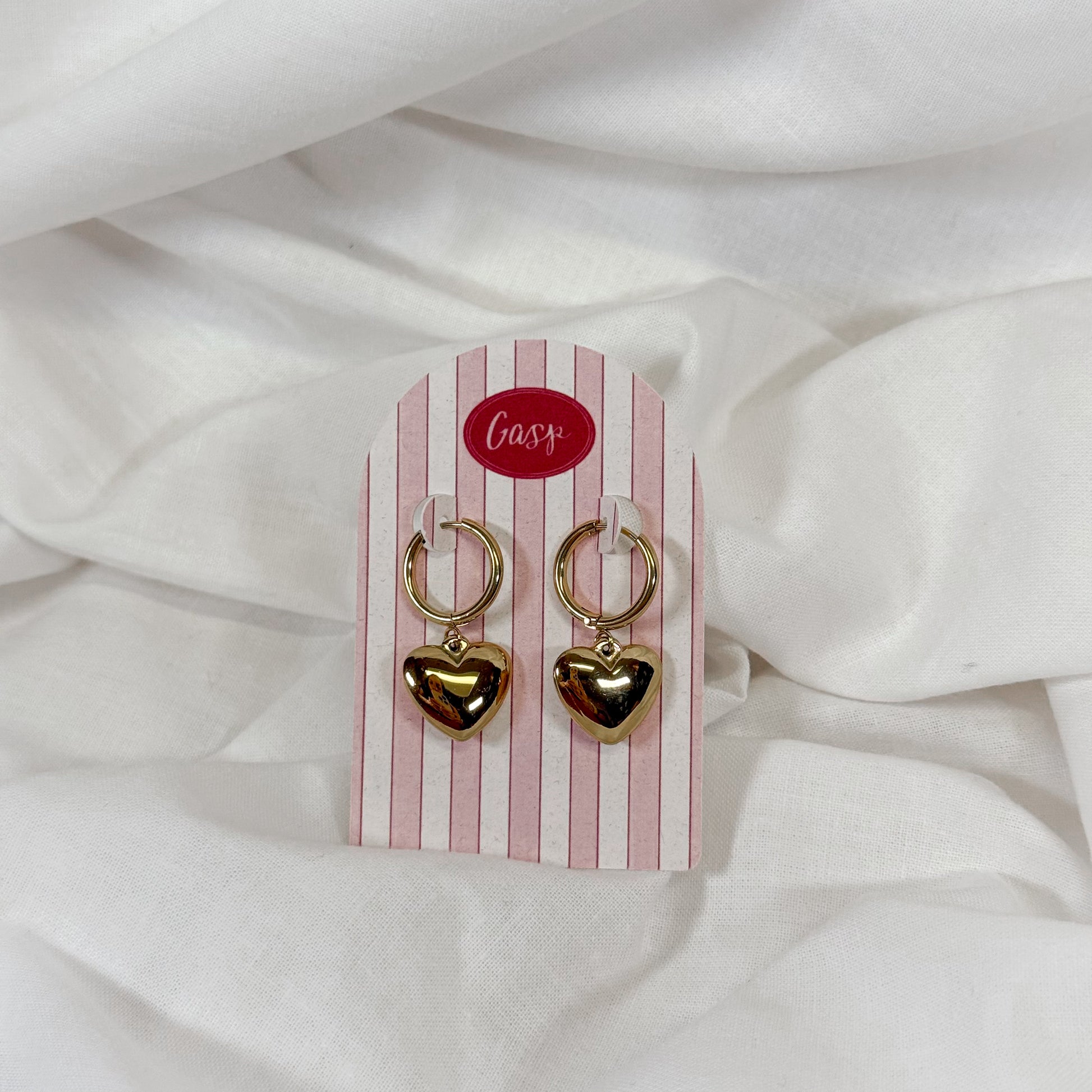 Puffy gold heart-shaped earrings on a pink and white striped card with 'Gasp' brand name, against a white fabric background.